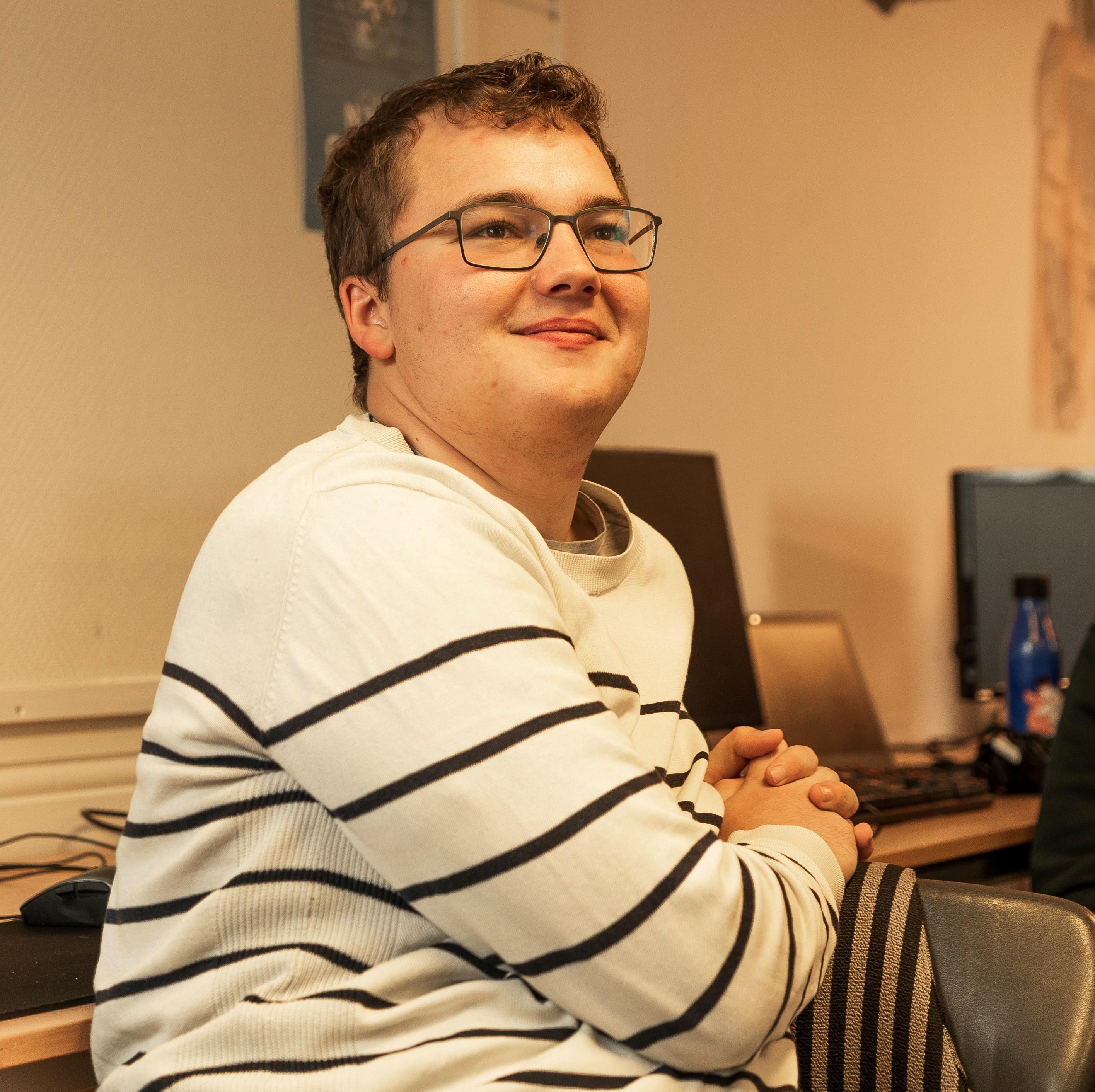 Ung mann med briller og stripete genser, sitter og ser til høyre for kamera