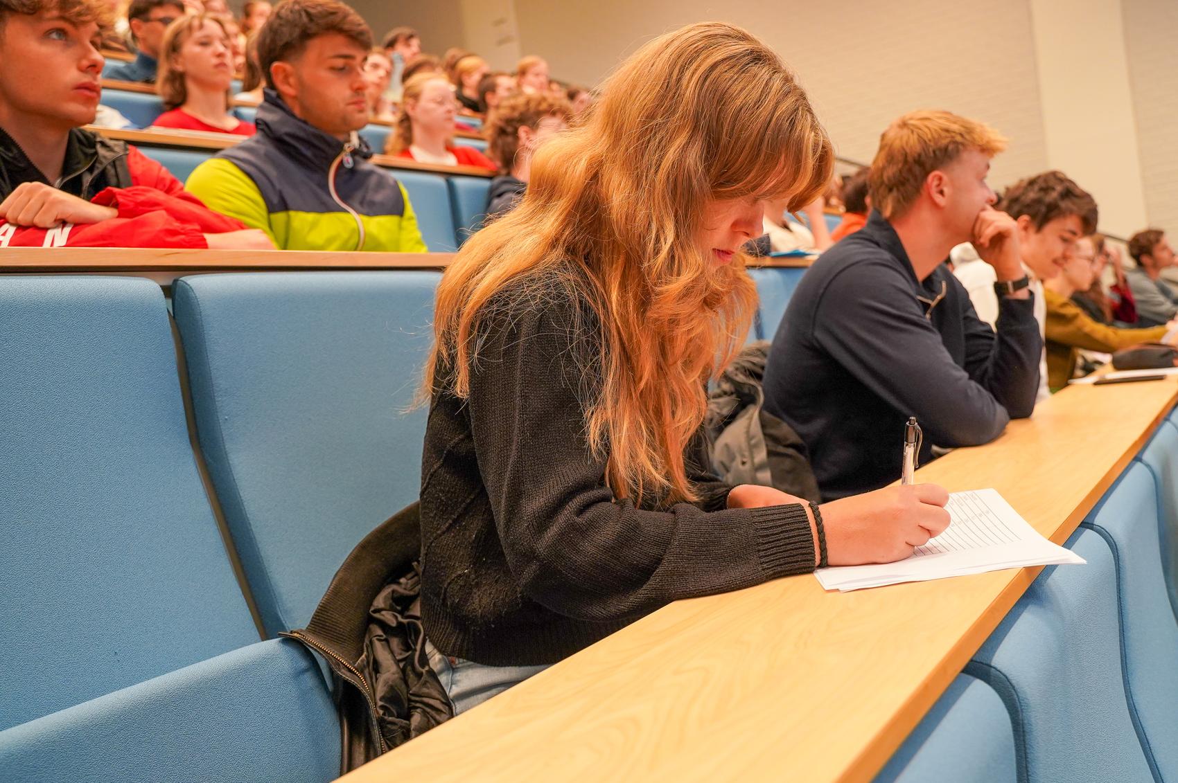 Jente sitter i auditorium fullt av folk og skriver på et ark
